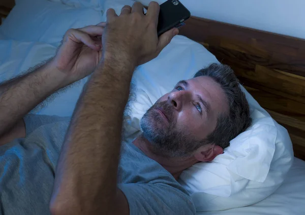 Junger attraktiver und entspannter Mann mit blauen Augen, der spät in der Nacht im Bett liegt, in dunklem und schummrigem Licht, Mobiltelefon oder Online-Dating in Internetsucht — Stockfoto