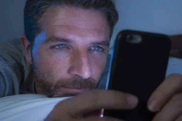 Joven hombre atractivo y relajado con los ojos azules acostado en la cama tarde en la noche en la red de luz oscura y tenue en el teléfono móvil o en línea citas en la adicción a Internet —  Fotos de Stock