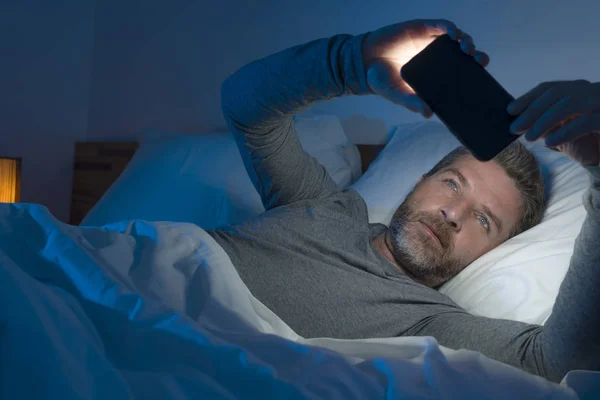 Giovane uomo attraente e rilassato con gli occhi azzurri sdraiato sul letto a tarda notte in rete luce scura e fioca sul telefono cellulare o in linea incontri in dipendenza da internet — Foto Stock