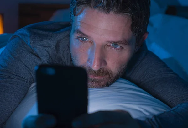 파란 눈을 가진 매력적 이고 편안 한 젊은 남자는 늦은 밤어두운 날씨에 휴대 전화로 또는 인터넷 중독으로 온라인 데이트를 하며 침대에 누워 있다. — 스톡 사진