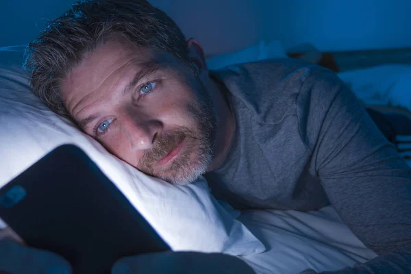 Young attractive and relaxed man with blue eyes lying on bed late at night in dark and dim light networking on mobile phone or online dating in internet addiction — Stock Photo, Image