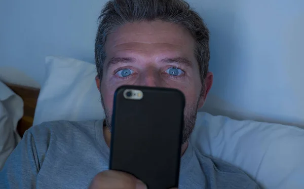 Atraente e louco viciado em internet homem com olhos azuis deitado na cama tarde da noite em rede de luz escura e escura no telefone celular ou on-line namoro em vício em mídia social — Fotografia de Stock