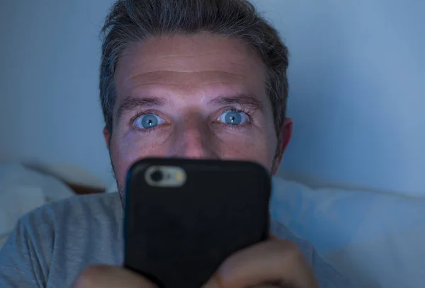 Привлекательный и сумасшедший интернет-наркоман с голубыми глазами, лежащий на кровати поздно ночью в темных и тусклых легких сетях на мобильном телефоне или онлайн знакомствах в социальных сетях — стоковое фото
