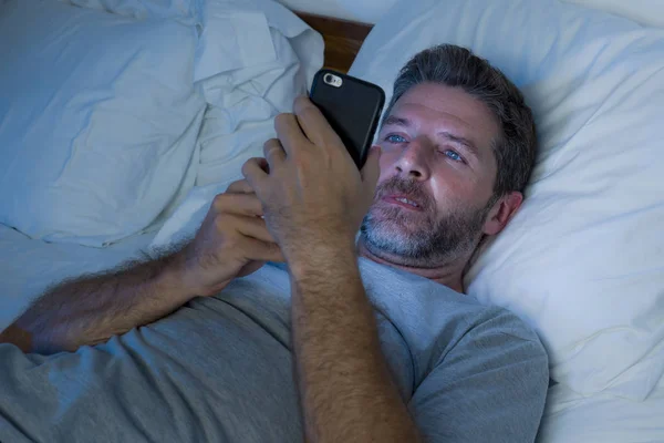 Happy man with blue eyes lying on bed late at night in dark light networking on mobile phone or online dating smiling relaxed enjoying internet in social media addiction — Stock Photo, Image