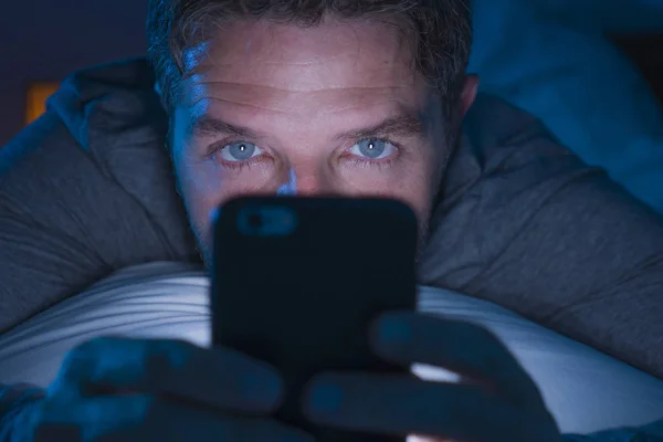 Привлекательный и сумасшедший интернет-наркоман с голубыми глазами, лежащий на кровати поздно ночью в темных и тусклых легких сетях на мобильном телефоне или онлайн знакомствах в социальных сетях — стоковое фото