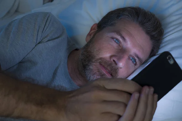Homem feliz com olhos azuis deitado na cama tarde da noite em rede de luz escura no telefone celular ou on-line namoro sorrindo relaxado curtindo internet no vício em mídia social — Fotografia de Stock