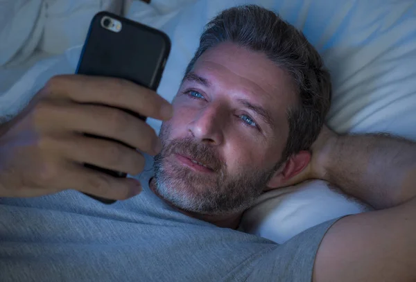 Glücklicher Mann mit blauen Augen, der spät in der Nacht im dunklen Licht auf dem Bett liegt, Netzwerkarbeit am Handy oder Online-Dating, entspannt lächelnd, Internet in Social-Media-Sucht genießend — Stockfoto