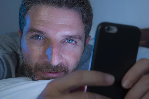 Homme heureux avec les yeux bleus couché sur le lit tard dans la nuit dans la lumière sombre réseautage sur téléphone mobile ou en ligne sortir ensemble sourire détendu profiter d "Internet dans la dépendance aux médias sociaux — Photo