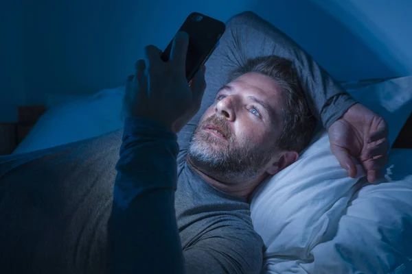 Glücklicher Mann mit blauen Augen, der spät in der Nacht im dunklen Licht auf dem Bett liegt, Netzwerkarbeit am Handy oder Online-Dating, entspannt lächelnd, Internet in Social-Media-Sucht genießend — Stockfoto