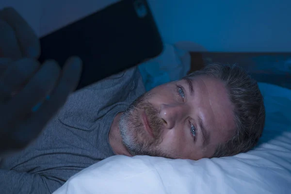 Glücklicher Mann mit blauen Augen, der spät in der Nacht im dunklen Licht auf dem Bett liegt, Netzwerkarbeit am Handy oder Online-Dating, entspannt lächelnd, Internet in Social-Media-Sucht genießend — Stockfoto