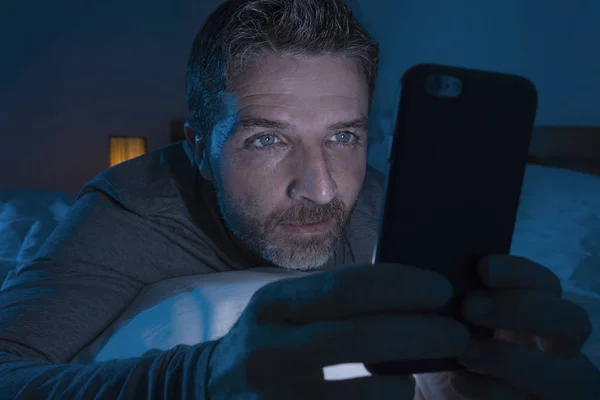 Happy man with blue eyes lying on bed late at night in dark light networking on mobile phone or online dating smiling relaxed enjoying internet in social media addiction — Stock Photo, Image