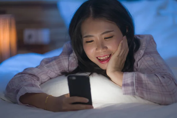 young beautiful and happy sweet Asian Chinese woman with in pajamas enjoying with mobile phone app in bed at night in internet social media addiction concept