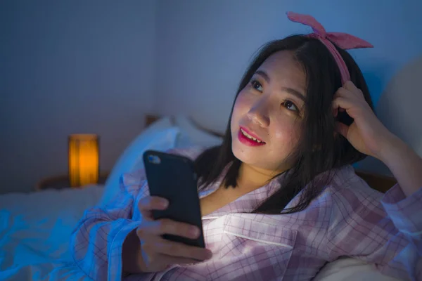 young beautiful and happy sweet Asian Korean woman with bow headband and pajamas networking with mobile phone in bed at night checking internet social media app