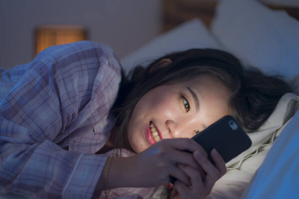 young beautiful and happy sweet Asian Korean woman with in pajamas enjoying with mobile phone app in bed at night in internet social media addiction concept