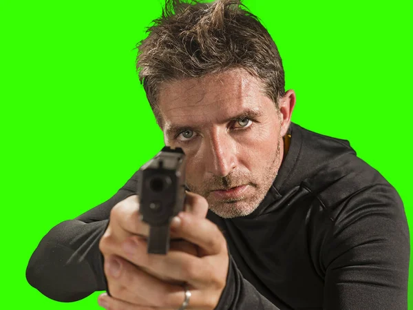 Chroma key green background action portrait of serious and attractive hitman or special agent man holding gun pointing the handgun to the camera on cinematic light — Stock Photo, Image