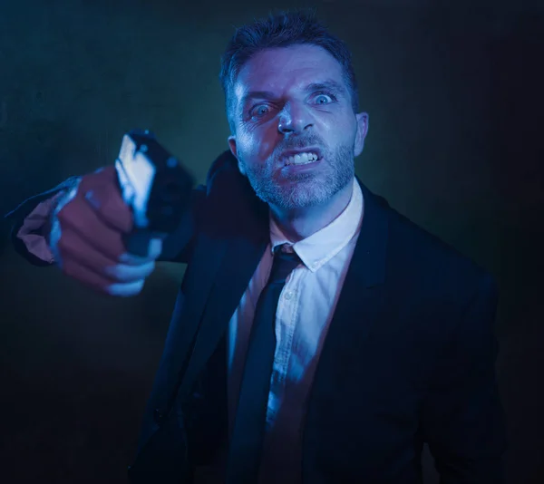 Scary dark portrait of young crazy psycho and furious man in businessman suit holding handgun in sick upset and threatening face expression isolated on grunge background — Stock Photo, Image