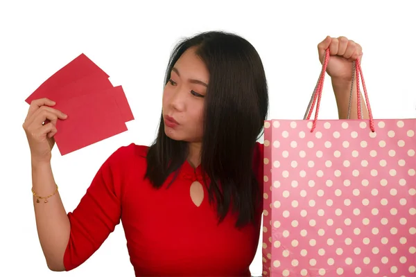 Młody szczęśliwy i piękny Azji dziewczyna w czerwonej sukience uśmiechnięty wesoły trzymając torby na zakupy jako podekscytowany wydawanie pieniędzy po otrzymaniu chiński Nowy Rok czerwona kieszeń z pieniędzy odizolowanych — Zdjęcie stockowe