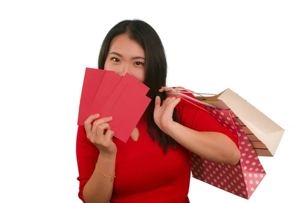 Młody szczęśliwy i piękny Azji dziewczyna w czerwonej sukience uśmiechnięty wesoły trzymając torby na zakupy jako podekscytowany wydawanie pieniędzy po otrzymaniu chiński Nowy Rok czerwona kieszeń z pieniędzy odizolowanych — Zdjęcie stockowe