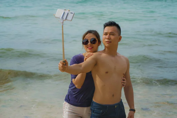 Sommar semester livsstil porträtt av unga glada och lekfulla asiatiska kinesiska par njuter på stranden tar stick selfie foto med handtelefon njuter av havet tillsammans — Stockfoto