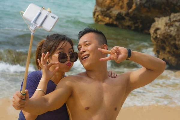 Summer holidays lifestyle portrait of young happy and playful Asian Chinese couple enjoying at the beach taking stick selfie photo with hand phone enjoying sea together — 스톡 사진