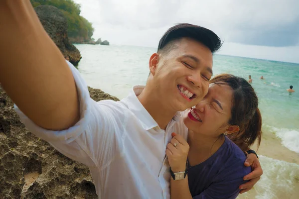 Summer holidays lifestyle portrait of young happy and playful Asian Chinese couple tourist enjoying at the beach taking selfie photo with hand phone enjoying sea together — 스톡 사진