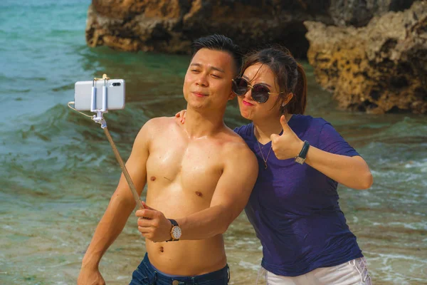 Summer holidays lifestyle portrait of young happy and playful Asian Chinese couple enjoying at the beach taking stick selfie photo with hand phone enjoying sea together — 스톡 사진