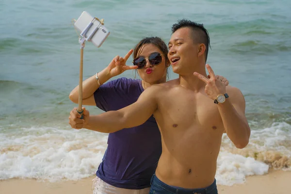Summer holidays lifestyle portrait of young happy and playful Asian Korean couple enjoying at the beach taking stick selfie photo with hand phone enjoying sea together — 스톡 사진