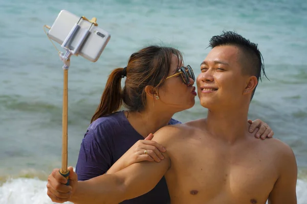 Sommar semester livsstil porträtt av unga glada och lekfulla asiatiska koreanska par njuter på stranden tar stick selfie foto med handtelefon njuter av havet tillsammans — Stockfoto