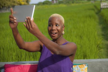 Çekici ve mutlu siyah Afro-Amerikan kadın havalı hipster tarzında özçekim yapıyor dışarıda dijital tabletle pirinç tarlasında yaz tatilinin tadını çıkarıyor.