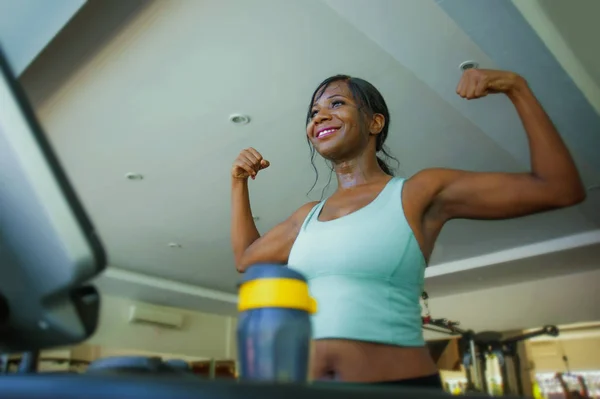 Νεαρή ελκυστική και ευτυχισμένη μαύρο afro αμερικανική γυναίκα κατάρτισης στο γυμναστήριο κάνει διάδρομο προπόνηση γυμναστήριο ποζάρουν παιχνιδιάρικο δείχνει μυς δικέφαλους σε αθλητικό σύλλογο χαμογελώντας χαρούμενα — Φωτογραφία Αρχείου