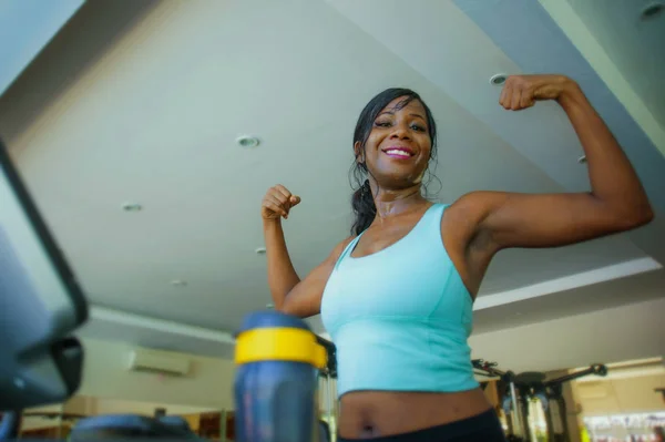 Joven atractivo y feliz negro afro americano mujer entrenamiento en gimnasio haciendo cinta de correr entrenamiento fitness máquina posando juguetón mostrando bíceps músculo en deportivo club sonriendo alegre — Foto de Stock