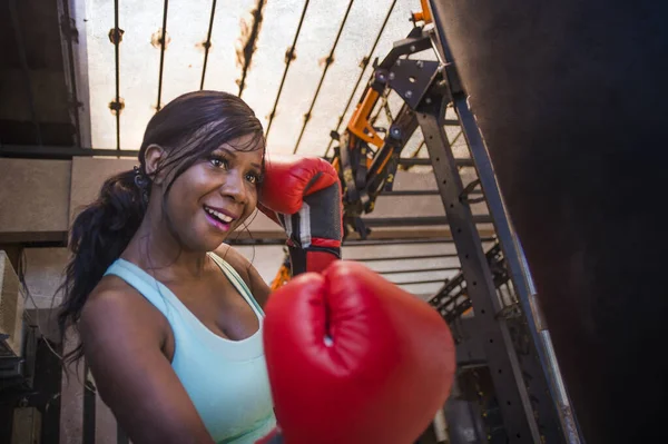 Gimnasio estilo de vida retrato de la joven atractiva y hermosa mujer afroamericana negro haciendo ejercicio en el gimnasio de entrenamiento sudoroso en la bolsa pesada puñetazos feliz con guantes de boxeo — Foto de Stock