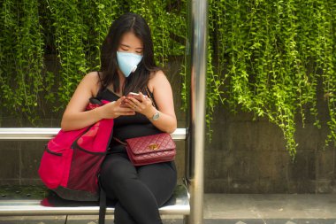 Korunaklı yüz maskesi takan Asyalı güzel ve çekici bir turist kadın otobüs durağında Coronavirüs salgını için cep telefonlarını kontrol ediyor.