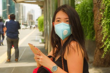 Korunaklı yüz maskesi takan Asyalı güzel ve çekici bir turist kadın otobüs durağında Coronavirüs salgını için cep telefonlarını kontrol ediyor.