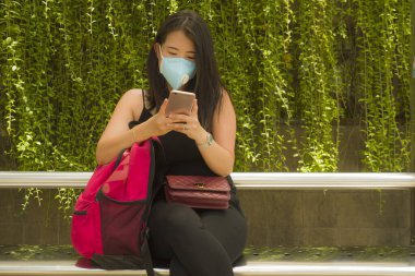 Korunaklı yüz maskesi takan Asyalı güzel ve çekici bir turist kadın otobüs durağında Coronavirüs salgını için cep telefonlarını kontrol ediyor.