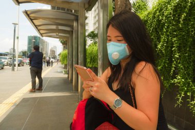 Korunaklı yüz maskesi takan Asyalı güzel ve çekici bir turistin portresi. Otobüs durağında Coronavirus salgını hakkında cep telefonuna bakıyor.