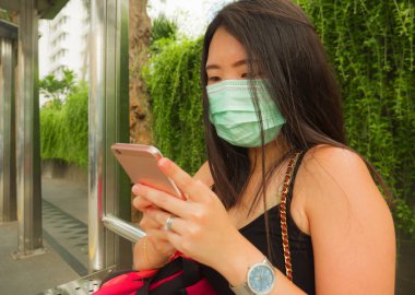 Korunaklı yüz maskesi takan Asyalı güzel ve çekici bir turist kadın otobüs durağında Coronavirüs salgını için cep telefonlarını kontrol ediyor.