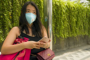 Korunaklı yüz maskesi takan güzel ve çekici Asyalı turist kadın otobüs durağında Coronavirüs salgını hakkında el telefonuna bakıyor.