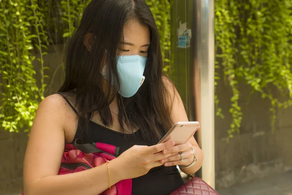 young beautiful and attractive Asian Chinese tourist woman wearing protective face mask checking hand phone at city bus stop about coronavirus outbreak