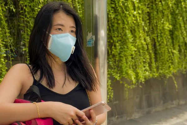 Joven hermosa y atractiva mujer turista china asiática con máscara facial protectora comprobando teléfono de la mano en la parada de autobús de la ciudad sobre el brote de coronavirus —  Fotos de Stock