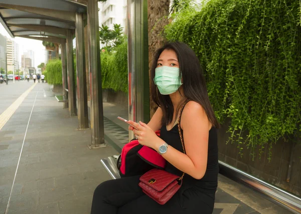 Якась вродлива і приваблива китайська жінка, одягнена в захисну маску, на зупинці міського автобуса перевіряє ручний телефон через спалах коронавірусу. — стокове фото