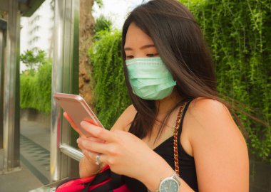 Korunaklı yüz maskesi takan güzel ve çekici Asyalı turist kadın otobüs durağında Coronavirüs salgını hakkında el telefonuna bakıyor.