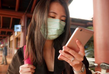 Genç ve güzel Asyalı Çinli kadın havaalanında koruyucu yüz maskesi takıyor Çin Coronavirus salgınında cep telefonuyla haberleri ve bilgileri kontrol ediyor.