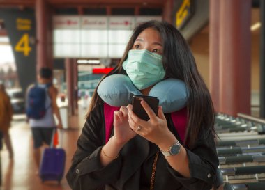 Genç ve güzel Asyalı Çinli kadın havaalanında koruyucu yüz maskesi takıyor Çin Coronavirus salgınında cep telefonuyla haberleri ve bilgileri kontrol ediyor.