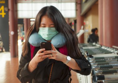 Genç ve güzel Asyalı Çinli kadın havaalanında koruyucu yüz maskesi takıyor Çin Coronavirus salgınında cep telefonuyla haberleri ve bilgileri kontrol ediyor.
