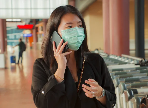 young beautiful Asian Chinese student woman at airport wearing protective facial mask talking on mobile phone on China Coronavirus epidemic outbreak spreading syndrome