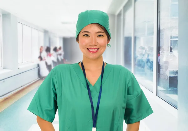 Young beautiful and happy Asian medicine doctor or chief nurse in green scrubs and surgeon hat smiling confident at hospital corridor background — Stock Photo, Image