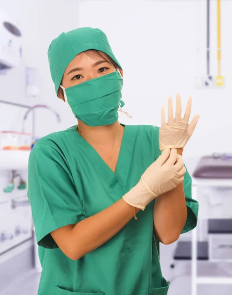 Молодая красивая и счастливая азиатская врач медицины или главная медсестра больницы в зеленых халатах и хирургической шляпе и маске положить латексные перчатки — стоковое фото