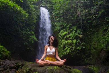 Genç çekici ve mutlu hippi kız dışarıda yoga yapıyor tropikal şelalede meditasyon yapıyor sağlıklı ve zen yaşam tarzında özgürlük ve doğanın tadını çıkarıyor.