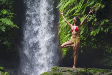 Genç çekici ve mutlu hippi kız dışarıda yoga yapıyor tropikal şelalede meditasyon yapıyor sağlıklı ve zen yaşam tarzında özgürlük ve doğanın tadını çıkarıyor.